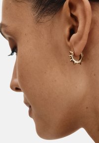 Guld ørering i hoop med spidse accenter, cirkulært design og tekstureret overflade, sat mod en neutral baggrund. Vist på en models øre.