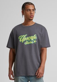 Grå oversized t-shirt med et farverigt "Upscale Studios" grafik med stjerner. Lavet af blødt stof, med korte ærmer og en afslappet pasform.