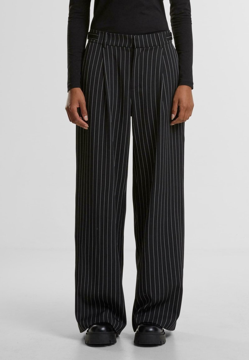 Personne portant un pantalon noir à jambes larges avec des fines rayures blanches verticales et des chaussures noires épaisses, debout devant un fond clair uni.