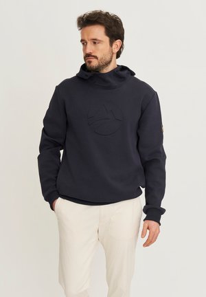 Man die een marineblauwe hoodie met reliëflogo en crèmekleurige broek draagt, staat met gedeeltelijk in de zakken gestoken handen en kijkt naar links.