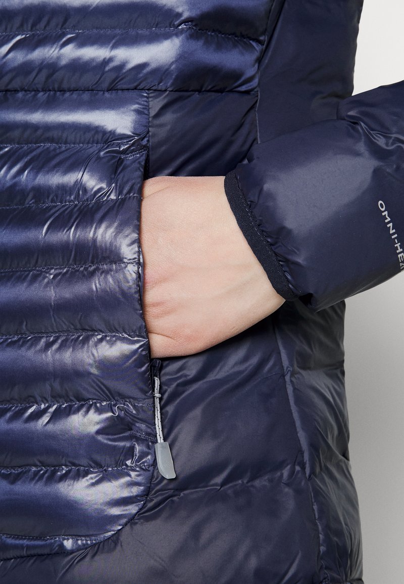 Main partiellement à l'intérieur de la poche zippée d'une veste matelassée bleu foncé avec une manche marquée "OMNI-HEAT".