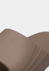 Lähikuva beigeistä liukusandalista, jossa on leveä, kuvioitu remmi ja muotoiltu pohja valkoisella taustalla.