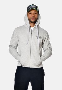Homme portant un sweat-zippé gris avec logo, un pantalon noir et une casquette camouflage verte Von Dutch, se tenant les mains dans les poches.