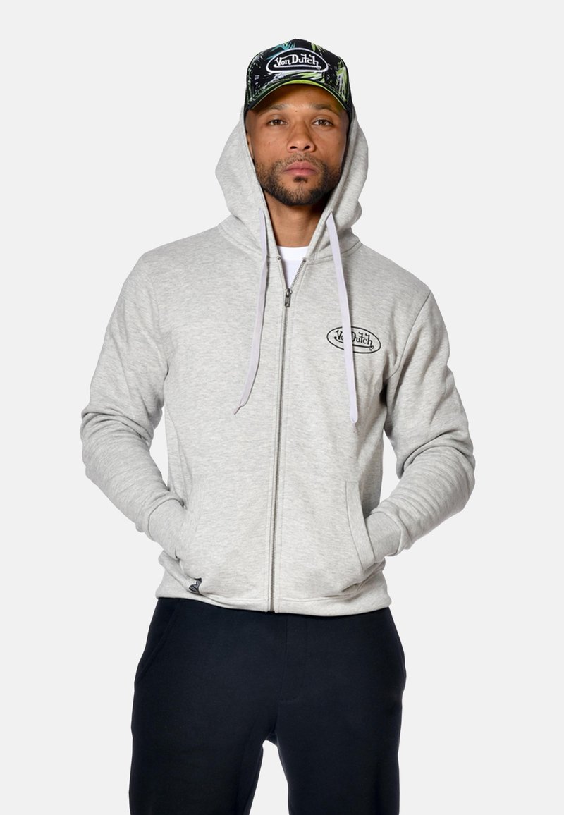 Homme portant un sweat-zippé gris avec logo, un pantalon noir et une casquette camouflage verte Von Dutch, se tenant les mains dans les poches.