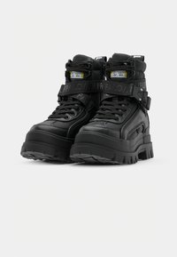 Black high-top boots made of synthetic leather, featuring prominent laces, a thick textured sole, and a Velcro strap with branding.