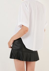 Weißes Kurzarmhemd mit abgerundetem Saum, kombiniert mit einem schwarzen, gefältelten Minirock aus Kunstleder. Das Outfit bietet eine glatte Textur und einen starken Kontrast.