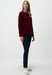 Pull en maille bordeaux à manches longues, associé à un pantalon slim bleu marine et des baskets blanches, sur un fond clair et uni.
