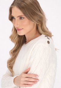 Maglione bianco a trecce con un motivo testurizzato, caratterizzato da tre bottoni metallici decorativi sulla spalla e un collo rotondo.