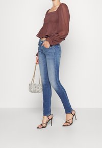 Blusa marrón de mangas abullonadas con escote cuadrado, jeans skinny azules, tacones negros de tiras y un pequeño bolso blanco con estampado.