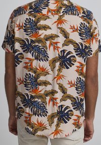 Camicia a maniche corte con sfondo pesca chiaro, caratterizzata da grandi motivi di foglie e fiori tropicali nelle tonalità blu navy, arancione e beige.