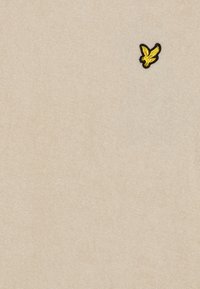 Μπεζ πλεκτό πουλόβερ φτιαγμένο από μαλακό ύφασμα, με μικρό μαύρο και κίτρινο κεντητό λογότυπο ενός πουλιού στην αριστερή περιοχή του στήθους.