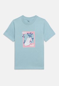 Lyseblå t-shirt med korte ærmer og et motiv af en hvid sneaker omgivet af blå blomster inde i en lyserød firkant, der viser Puma-logoet.