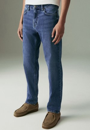 Személy kék, egyenes szárú farmernadrágot és barna, velúr fűzős cipőt visel, semleges háttér előtt áll.