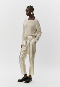 Bežinė megztinis, blizgūs auksinės spalvos kelnės iki juosmens su tekstūruotu paviršiumi ir juodi loaferiai. Modelis stovi tiesiai, dėvėdama dideles apvalias auskarus.