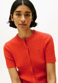 Roter Strickcardigan mit einem strukturierten Rautenmuster, kurzen Ärmeln und Knöpfen vorne. Kleiner Logopatch auf der linken Brust platziert.