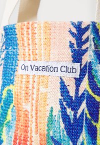 Szövetből készült bevásárlótáska élénk színekkel, csíkos és levélmintás dizájnnal, rajta "On Vacation Club" feliratú textilcímkével.