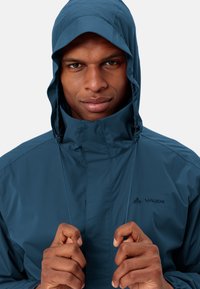 Blå vattentät jacka med full huva, slät textur, hög krage och logodetalj. Har en strömlinjeformad design och justerbara stängningar.