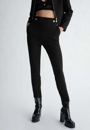 Mujer que lleva pantalones negros de talle alto con botones dorados, chaqueta negra recortada, blusa negra de encaje transparente y botas negras de plataforma brillantes.