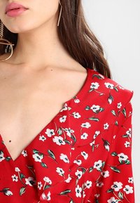 Vestido floral rojo con mangas cortas, que presenta un escote en V. La tela muestra flores blancas y hojas verdes sobre una textura suave.
