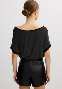 Top negro de corte holgado con textura brillante y diseño tipo off-the-shoulder; combinado con pantalones cortos negros elegantes, que muestran un material suave y líneas limpias.