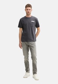 Camiseta de manga corta gris oscuro con un bolsillo en el pecho, combinada con jeans gris claro y zapatillas blancas. Diseño simple con una textura sutil.