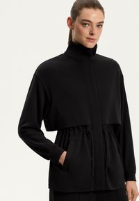 Zwarte cropped jas met hoge kraag, elastische taille met koord, zijzakken en lange mouwen. Glad textuur en minimalistisch ontwerp.