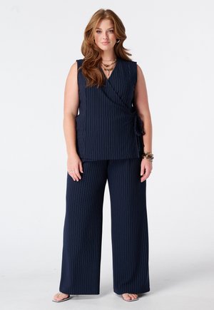 Vrouw met lichtbruin haar die een marineblauw gestreept mouwloos overslagtopje en bijpassende broek met wijde pijpen draagt, staand op een witte achtergrond.