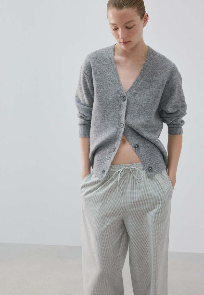 Femme avec les mains dans les poches du pantalon, portant un cardigan gris ample boutonné et un pantalon gris clair à cordon, sur un fond uni.