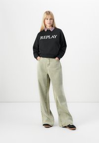Vrouw staat met handen in de zakken, draagt een zwart "REPLAY" sweatshirt over een geblokte kraagshirt, brede lichtgroene jeans en zwarte sneakers.