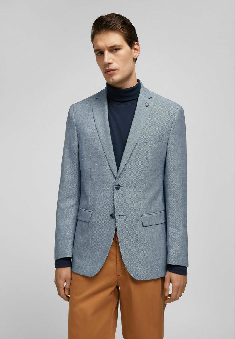 Lichtblauwe textuurblazer met notch-lapels, twee knopen en voorzakken, gedragen over een marineblauwe coltrui en gecombineerd met een bruine broek.