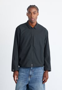 Sort zip-up jakke med krave, lange ærmer og en glat tekstur. Bæres over blå denim jeans med regular fit.
