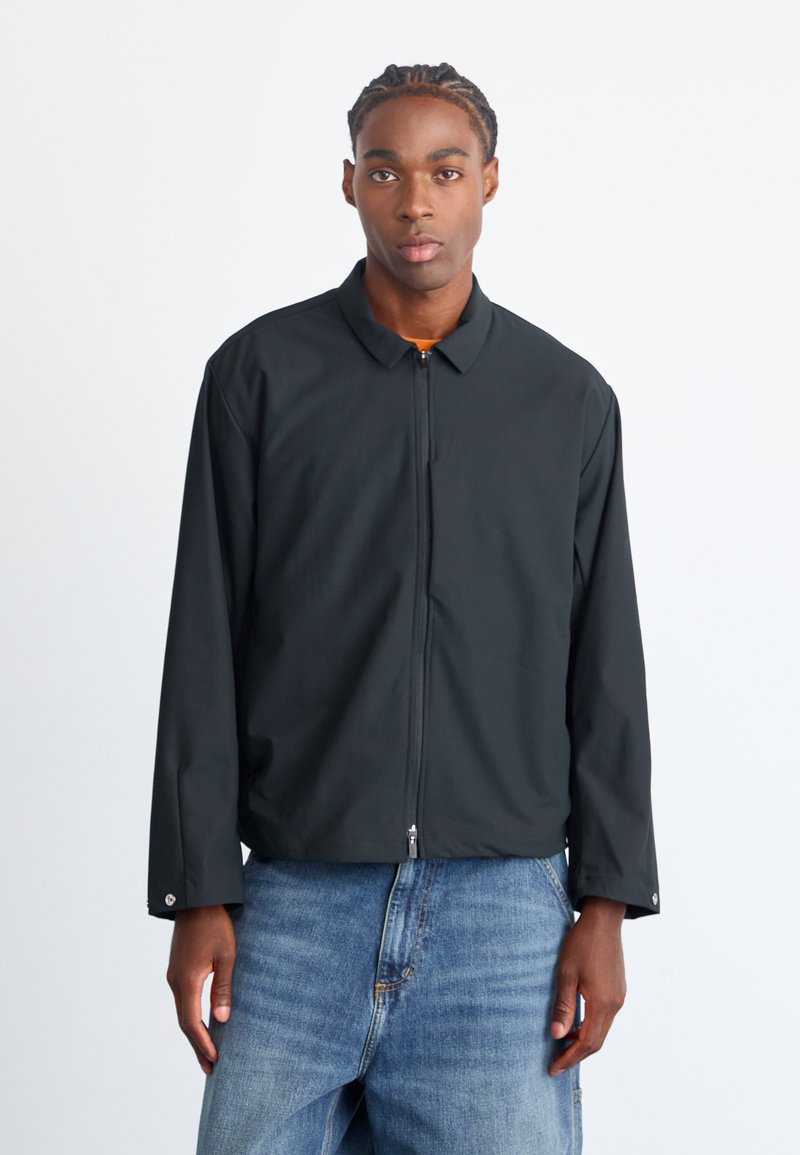 Sort zip-up jakke med krave, lange ærmer og en glat tekstur. Bæres over blå denim jeans med regular fit.