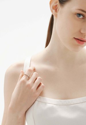 Joven con ojos azules y cabello castaño toca la tira de su hombro, llevando una blusa blanca y un anillo plateado en la mano izquierda.