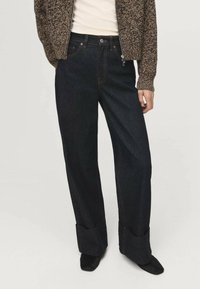 Vaqueros de mezclilla azul oscuro de pierna ancha con un ajuste ligeramente suelto y dobladillos doblados hacia arriba. Presentan costuras contrastantes en naranja y diseño estándar de cinco bolsillos.