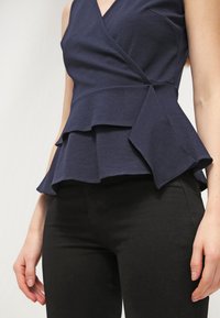 Námořnická modrá zavinovací blůza vyrobená z jemného materiálu, se zdobením ve vrstveném peplum designu a výstřihem do V. Kombinována s upnutými černými kalhotami.