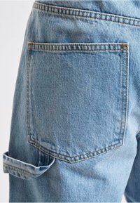 Lys blå denim jeans med en baglomme, har kontrasterende orange syninger og en oppekrøllet kant. Teksturen ser let falmet ud.