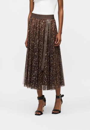 Žena v hnedom midi sukni s leopardím vzorom a skladaním, s tmavým pásom, bielom bezrukávovom topom a čiernych sandáloch na vysokom opätku s remienkami okolo členkov.