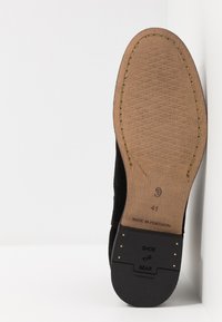 Chaussure en daim noir avec un bout arrondi, présentant une semelle en bois clair et un talon en caoutchouc noir. La partie inférieure texturée révèle des détails de marque et de taille.