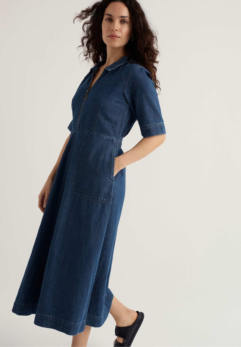 Frau mit lockigem Haar, die ein blaues Denim-Midikleid mit Vorder Taschen, halblangen Ärmeln, Kragen, Reißverschluss und schwarzen Pantoletten trägt.