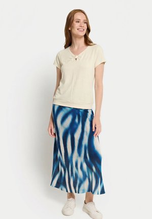 Lächelnde Frau, die ein beigefarbenes Kurzarmoberteil mit Knotendetail und einen blau gemusterten Midirock trägt, kombiniert mit weißen Turnschuhen.