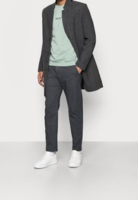 Abrigo de mezcla de lana gris, sudadera verde claro, pantalones gris oscuro y zapatillas blancas. El abrigo tiene un cuello alto y botones en la parte frontal.