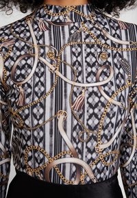 Svart och vit mönstrad topp med guldiga kedjor, fransar och band på en texturerad bakgrund. Figursydd design med hög hals.