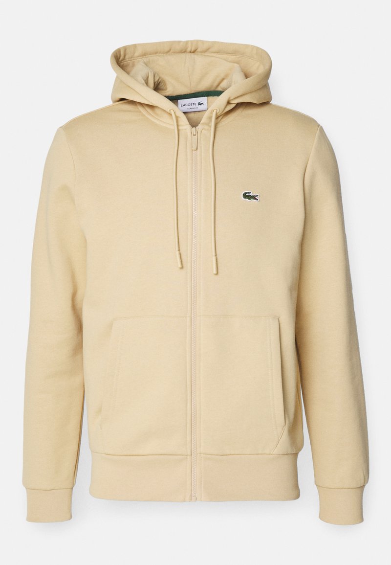 Beige Kapuzenjacke mit Reißverschluss aus weichem Material, ausgestattet mit einer Kängurutasche, Zugbändern und einem grün akzentuierten Logo auf der Brust.