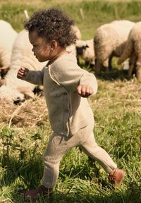 Un bambino in un outfit di maglina beige che cammina sull'erba vicino a diverse pecore che pascolano in un campo soleggiato.
