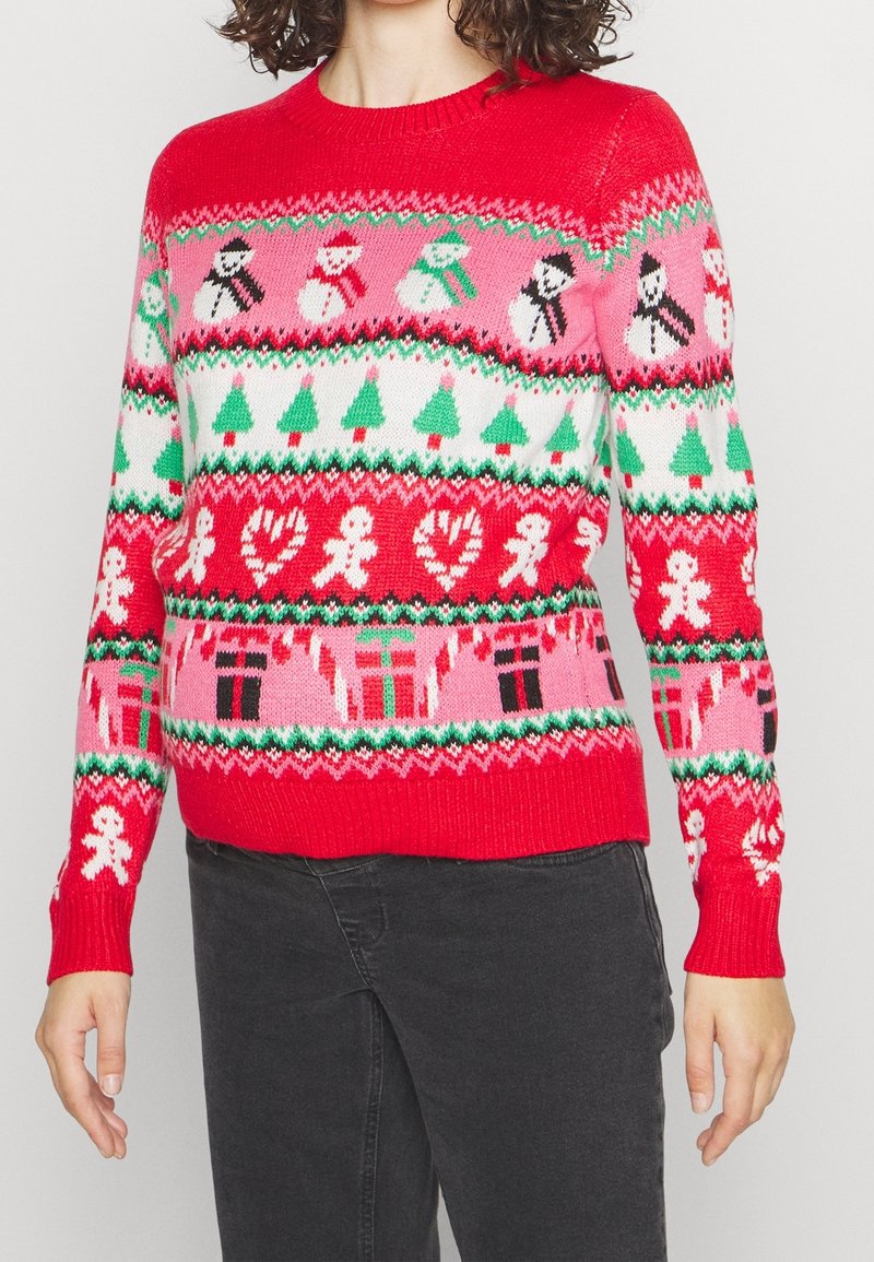 Pull de Noël rouge avec des bonhommes de neige, des sapins de Noël, des bonshommes en pain d'épice, des cannes de bonbon, des cœurs et des motifs de cadeaux, porté avec un pantalon foncé.