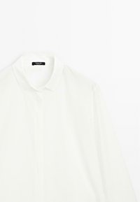 Camicia bianca a maniche lunghe con colletto classico, realizzata in tessuto liscio. Presenta una pattina dei bottoni nascosta e un design minimalista.