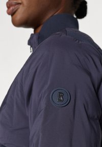 La veste isolée navy présente un tissu texturé, une fermeture éclair, un col montant et un écusson circulaire avec un « B » en relief sur la manche.