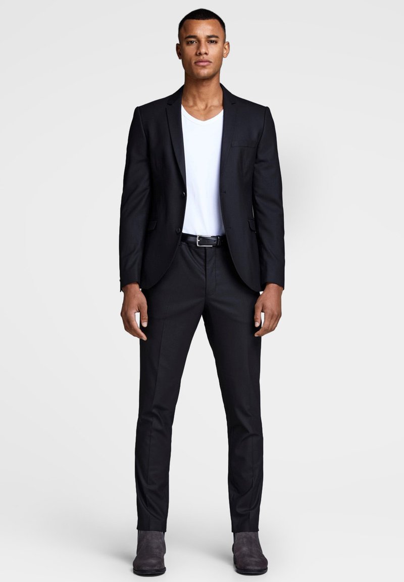 Black suit with a single button, paired with a white V-neck t-shirt. Slim-fit trousers and grey ankle boots complete the look.