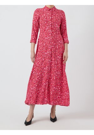 Woman wearing a red long-sleeve floral maxi dress with a collar and black pointed heels, standing against a plain background.