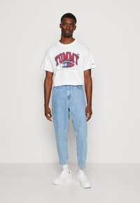 Tricou alb cu imprimeu roșu și albastru "TOMMY JEANS", asortat cu blugi deschiși la culoare, cu talie înaltă, și adidași albi. Fundal neutru.